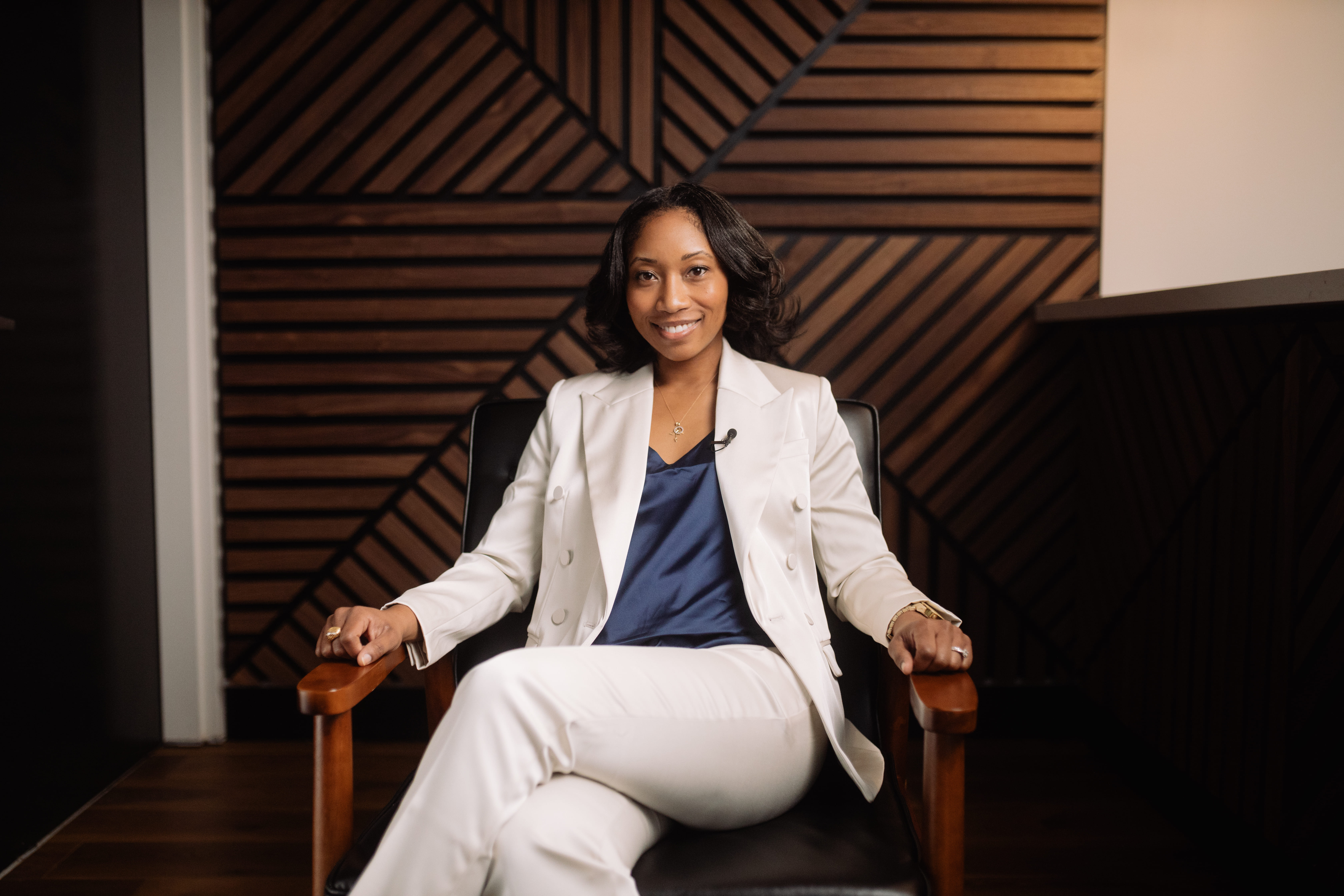 Dr. Scarlett sitting in a black leather armchair, wearing a white suit and navy top against a wooden accent wall.